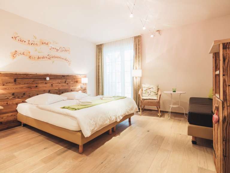 In einem gemütlichen Schlafzimmer mit Holzbettkopfende und Wanddekoration steht ein Bett, daneben ein Sessel und ein Beistelltisch, während rechts ein Teil eines Holzschranks und ein Sofa sichtbar sind.