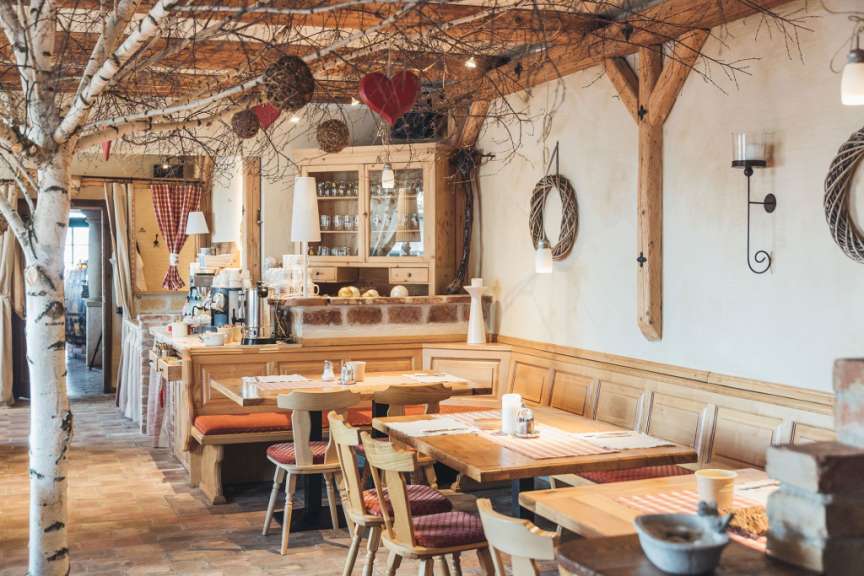 Das Bild zeigt einen rustikalen Restaurantbereich mit Holztischen, Sitzbänken und Wanddekorationen wie Kränzen und Herzen.