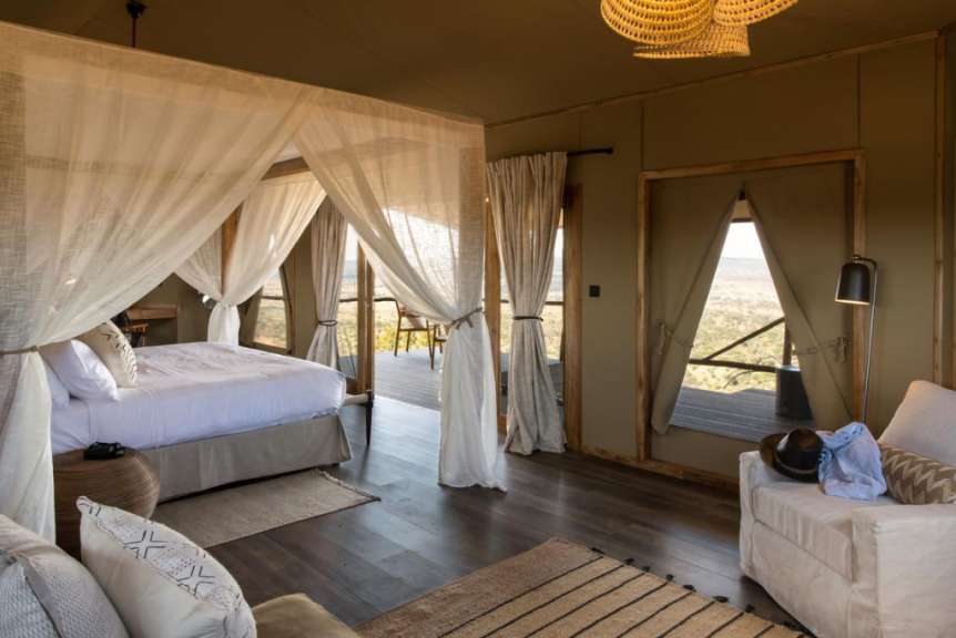 Ein luxuriöses Zeltzimmer mit einem Himmelbett, das auf eine Außenterrasse mit Blick auf eine Savannenlandschaft blickt.