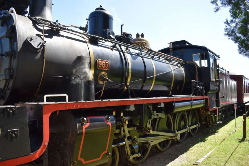Ein Dampfzug mit der Nummer 967 steht auf Schienen, mit Dampf, der aus einem Zylinder aufsteigt, und einem blauen Himmel im Hintergrund.