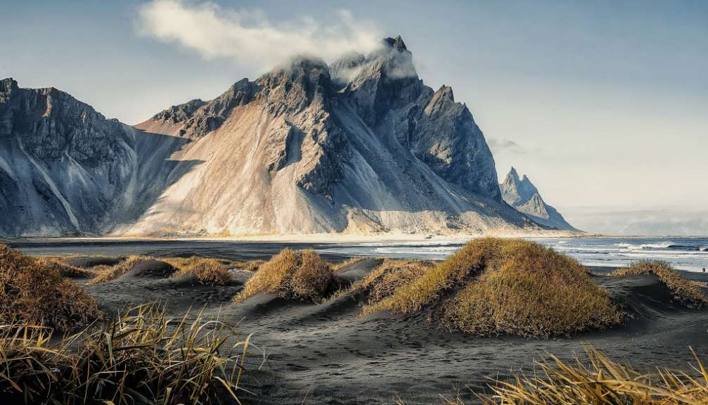 Eine zerklüftete Bergkette mit Wolken, die sich um die Gipfel wickeln, erhebt sich hinter einer Küstenlandschaft aus schwarzem Sand, Dünen mit goldenem Gras und dem tosenden Meer.