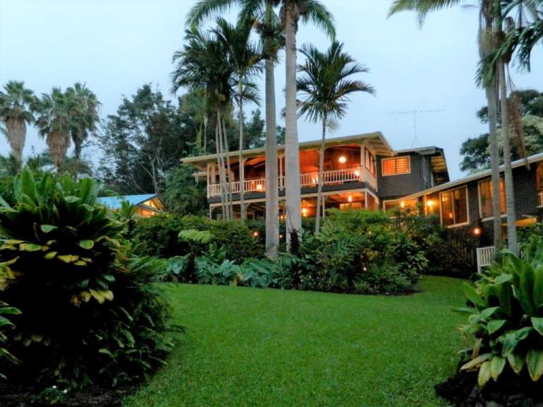Ein Haus mit einer Veranda, umgeben von üppiger Vegetation und Palmen, wird am Abend von den Lichtern beleuchtet.