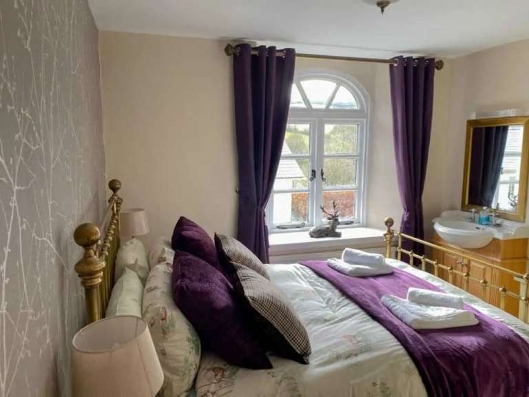 Ein Schlafzimmer mit einem goldenen Bettgestell, lila Vorhängen, einem Waschbecken und Blick aus dem Fenster.