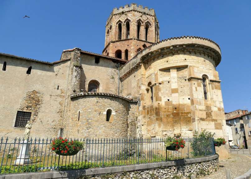 Ein großer, romanischer Glockenturm ragt über die Dächer eines Steingebäudes, das von einer schmiedeeisernen Absperrung mit Blumenkästen umgeben ist.