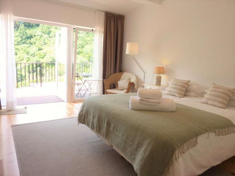 Ein Schlafzimmer mit Bett, Sessel, Nachttischen und einer Tür, die auf einen Balkon mit Blick ins Grüne führt.