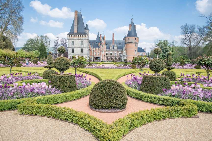 Eine Burg mit einem kunstvoll angelegten Garten voller Tulpen und Buchsbaumhecken unter blauem Himmel mit weißen Wolken.
