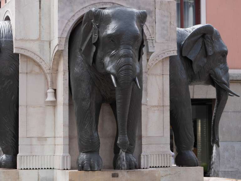Zwei Elefantenstatuen aus dunklem Metall stehen in einer steinernen Architektur, wobei Wasser aus dem Rüssel einer Statue spritzt.