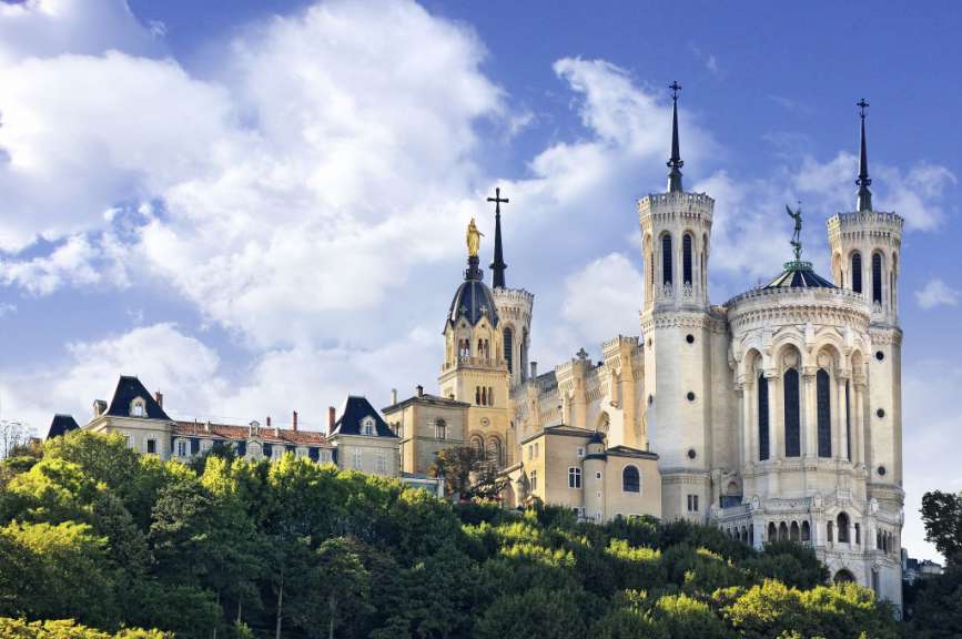 Die Basilika Notre-Dame de Fourvière thront auf einem Hügel über der Stadt Lyon, im Hintergrund ist ein blauer Himmel mit Wolken zu sehen.