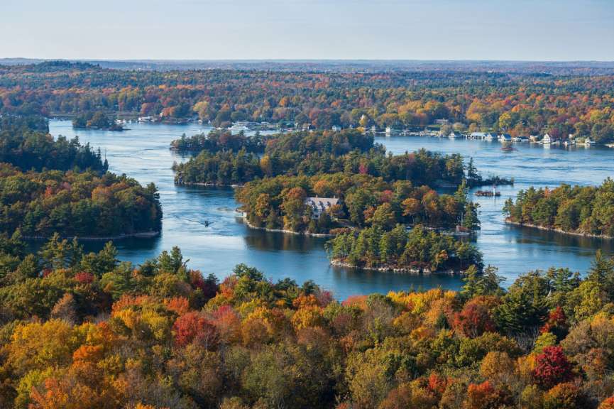 Ein Weitwinkelbild mit einer Drohne zeigt die Inseln der Thousand Islands im Herbst, wobei der Fluss von bunten Bäumen gesäumt ist.