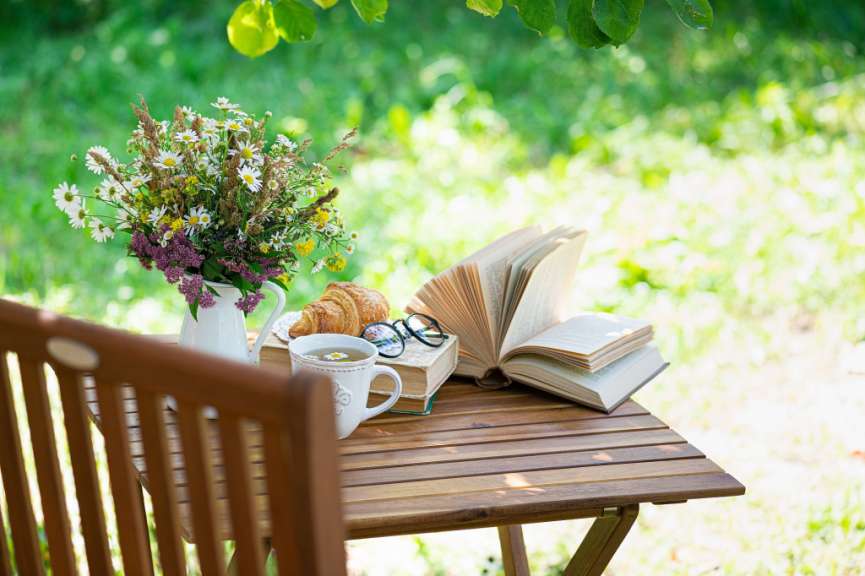 Ein Blumenstrauß, ein Croissant, eine Tasse Tee und Bücher liegen auf einem Holztisch im Freien.