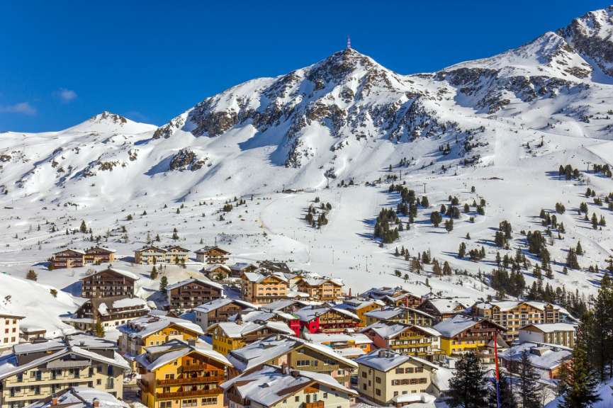 Ein Bergdorf mit bunten Häusern liegt im Schnee vor einer schneebedeckten Bergkette.