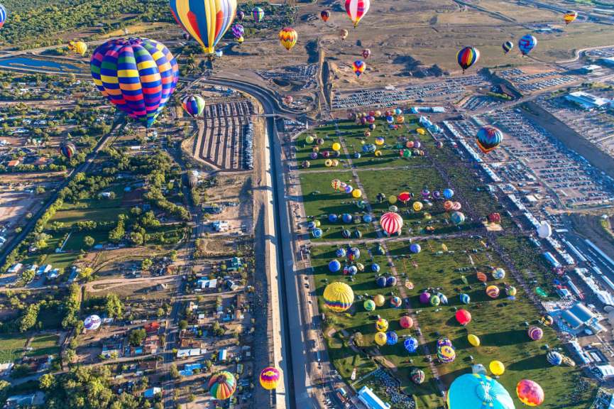 Eine riesige Anzahl von bunten Heißluftballons schwebt am Himmel über einem großen Gelände mit vielen parkenden Autos und Menschen, die auf dem grünen Rasen stehen.