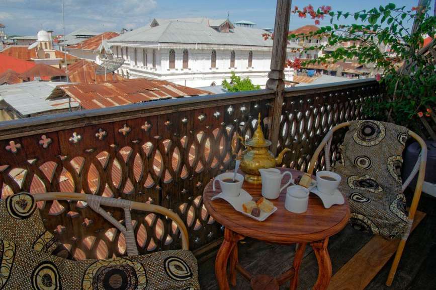 Auf einem Balkon mit Blick auf eine Stadt im Sonnenlicht stehen ein Tisch und ein Stuhl mit Kaffee und Gebäck.