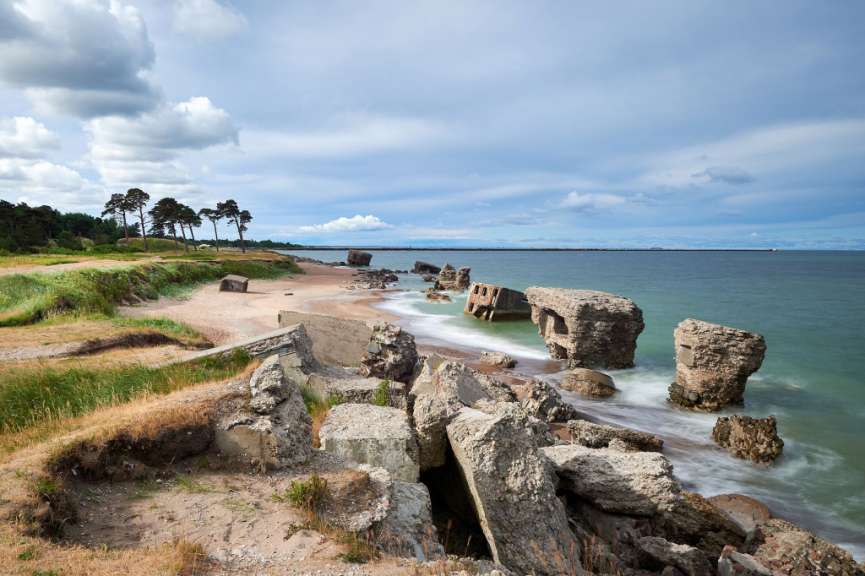 Ruinen von Betonstrukturen im Wasser und am Ufer eines sandigen Strandes unter einem bewölkten Himmel.