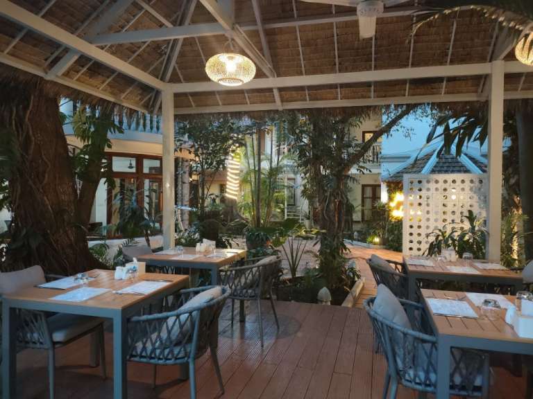 Der Innenhof eines Restaurants ist mit Holztischen und -stühlen möbliert, umgeben von üppiger Vegetation und mit einer Dachkonstruktion aus Stroh.