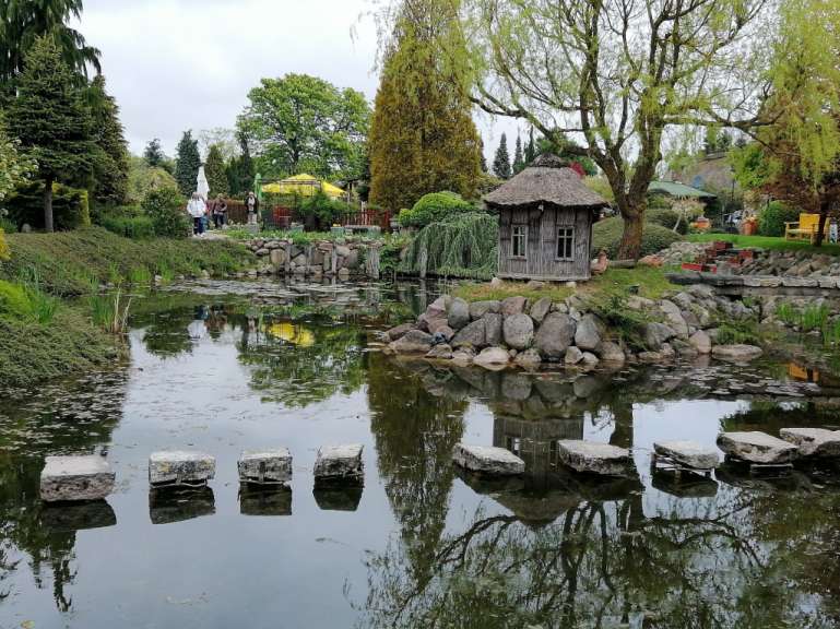 Ein kleiner Teich mit einer Reihe von Trittsteinen schlängelt sich durch einen üppigen Garten mit Bäumen, Sträuchern, einer kleinen Hütte mit Strohdach und Steinen.