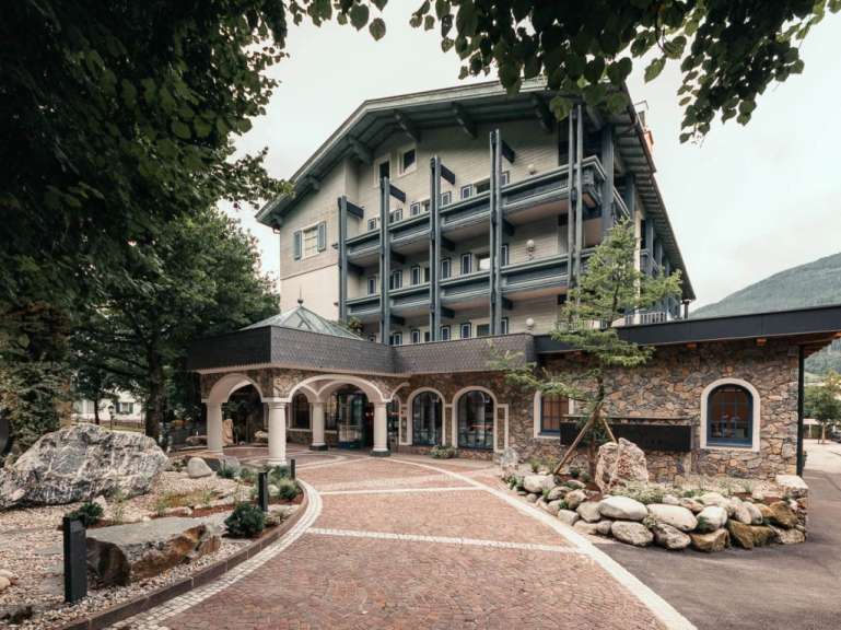 Die Fassade eines Hotels aus Stein und Holz mit vielen Balkonen, umgeben von Bäumen und einem gepflasterten Weg, ist zu sehen.