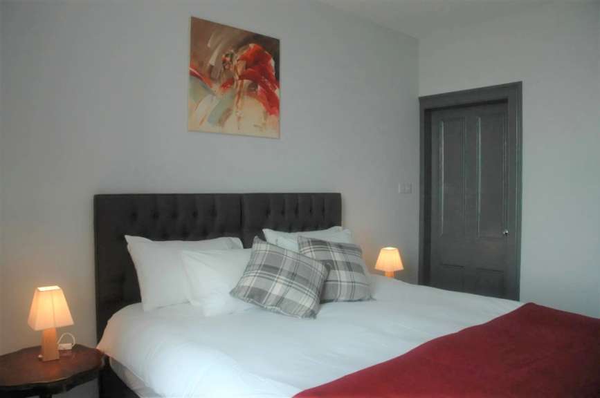 Ein Bett mit einem dunkelgrauen Kopfteil, weißen Laken und Kissen mit kariertem Muster, sowie einem roten Überwurf steht in einem Raum mit grauer Wand, einem Kunstwerk und einer Holztür.