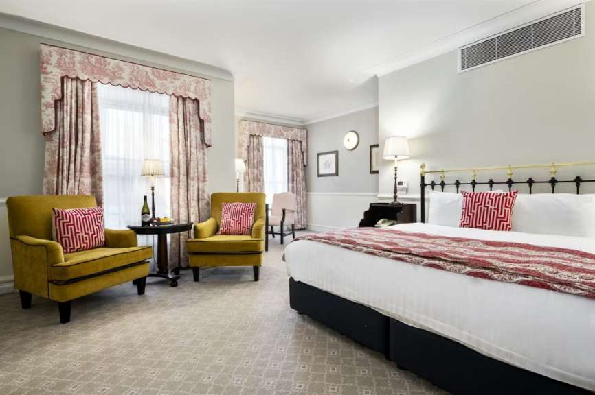 In einem Hotelzimmer stehen zwei gelbe Sessel mit roten Kissen vor verzierten Vorhängen und einer Fensterfront, während im Hintergrund ein Bett mit roter Tagesdecke und passenden Kissen zu sehen ist.