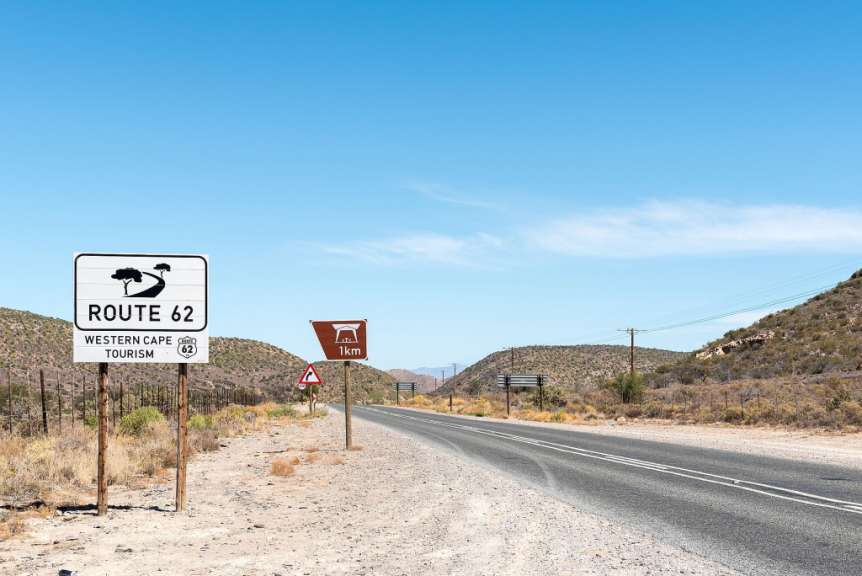 Auf einem Foto ist die Straße Route 62 mit Schildern im westkap-touristischen Stil zu sehen, die auf einem sonnigen Tag mit blauem Himmel vor einer trockenen, hügeligen Landschaft zu sehen ist.