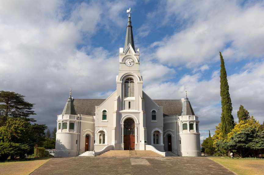 Eine Kirche mit einer hohen Turmspitze mit Uhr und Wetterhahn wird von einem Baum und Büschen auf der linken Seite und einem hohen, schlanken Baum auf der rechten Seite eingerahmt.