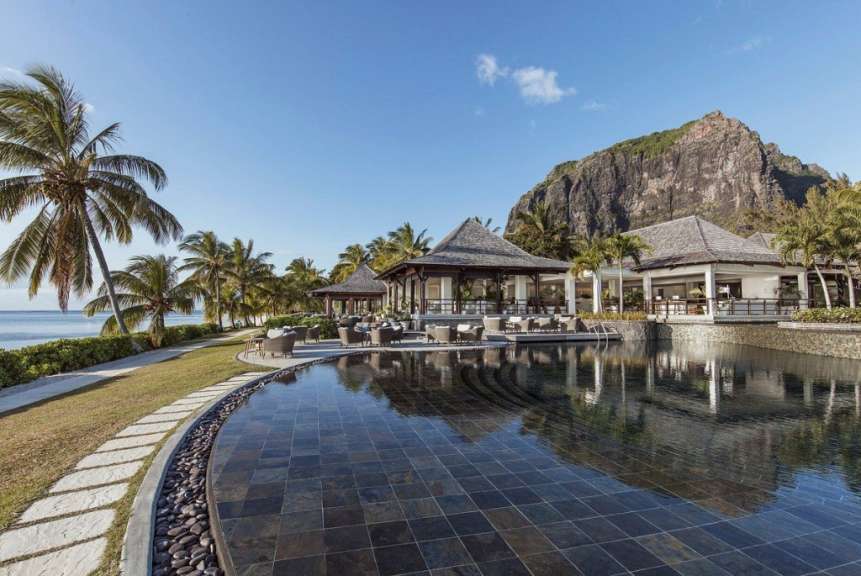 Ein Pool mit reflektierendem Wasser vor einem tropischen Resort mit Palmen und einem Berg im Hintergrund.