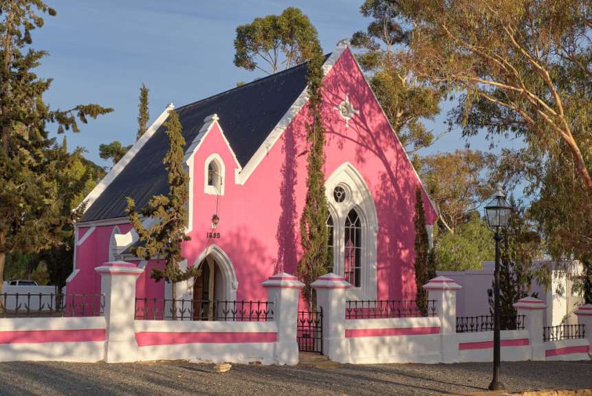 Eine pinke Kirche mit einem schwarzen Dach, umgeben von Bäumen und Zäunen.