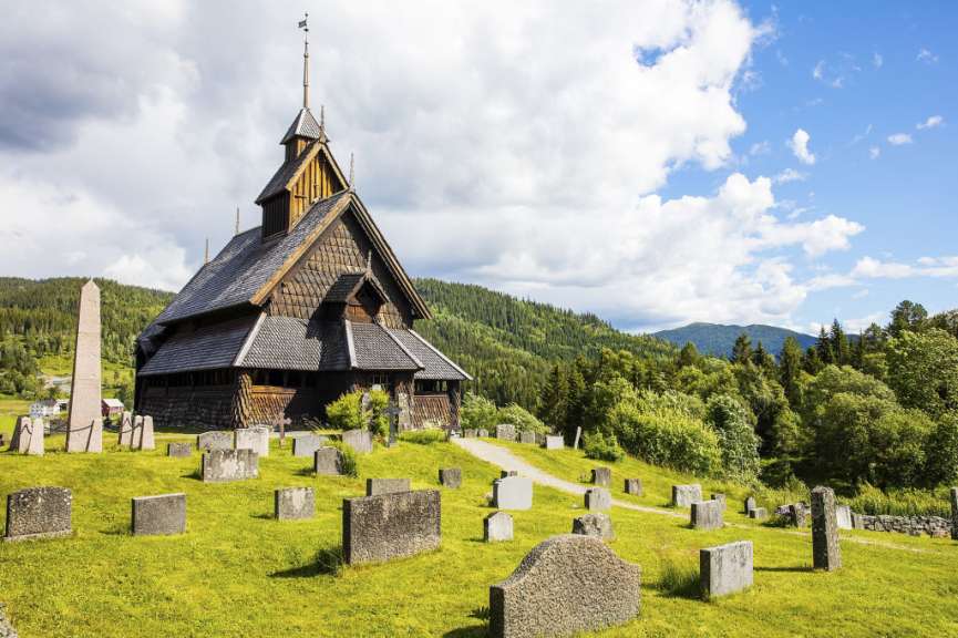 Ein hölzerner Stabkirchenturm mit einem Friedhof im Vordergrund und bewaldeten Hügeln im Hintergrund.