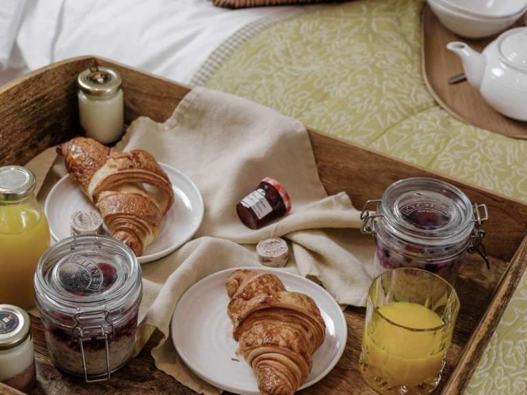 Auf einem hölzernen Tablett in einem Bett werden zwei Croissants mit Marmelade, Butter, Haferflocken und Orangensaft serviert.