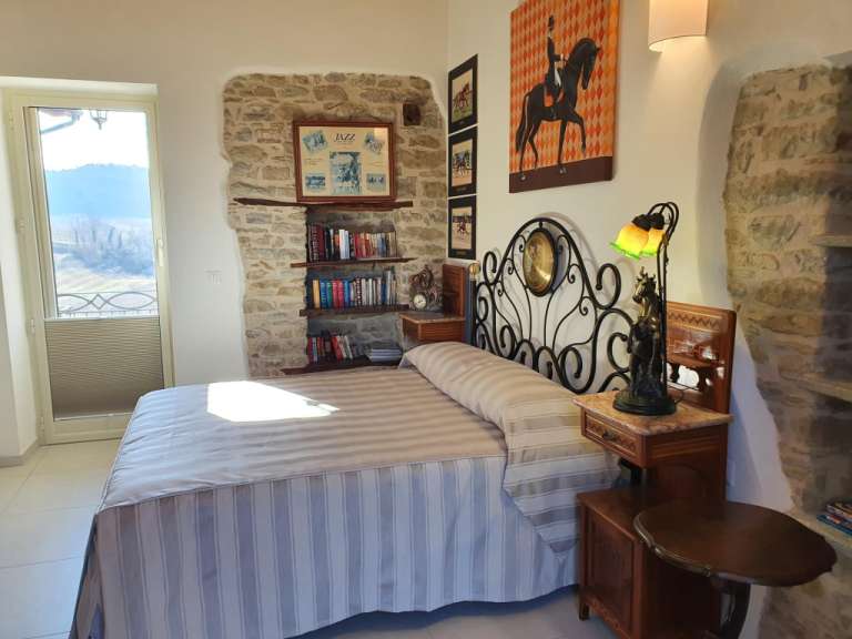 Ein Schlafzimmer mit einem Bett, das von einer Steinkonstruktion und Dekorationsgegenständen umgeben ist, wird von einem großen Fenster mit Blick auf eine ländliche Landschaft beleuchtet.