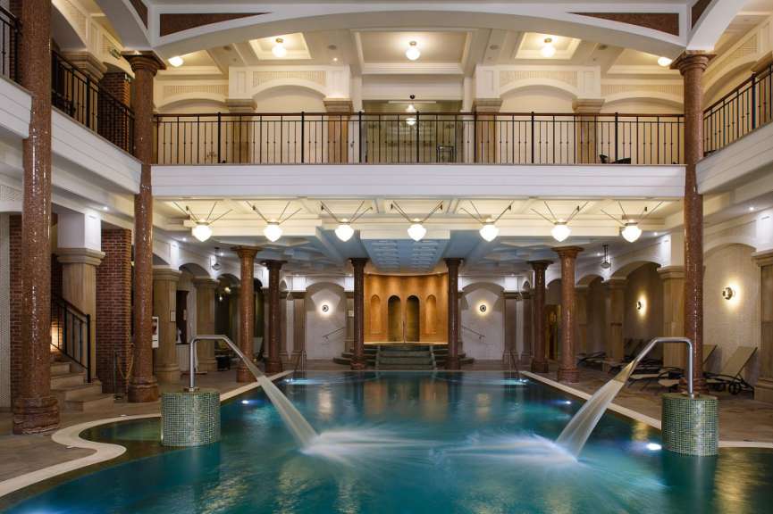 Ein Innenpool mit Wasserspielen und Liegestühlen wird von Säulen und einer Galerie umrahmt.