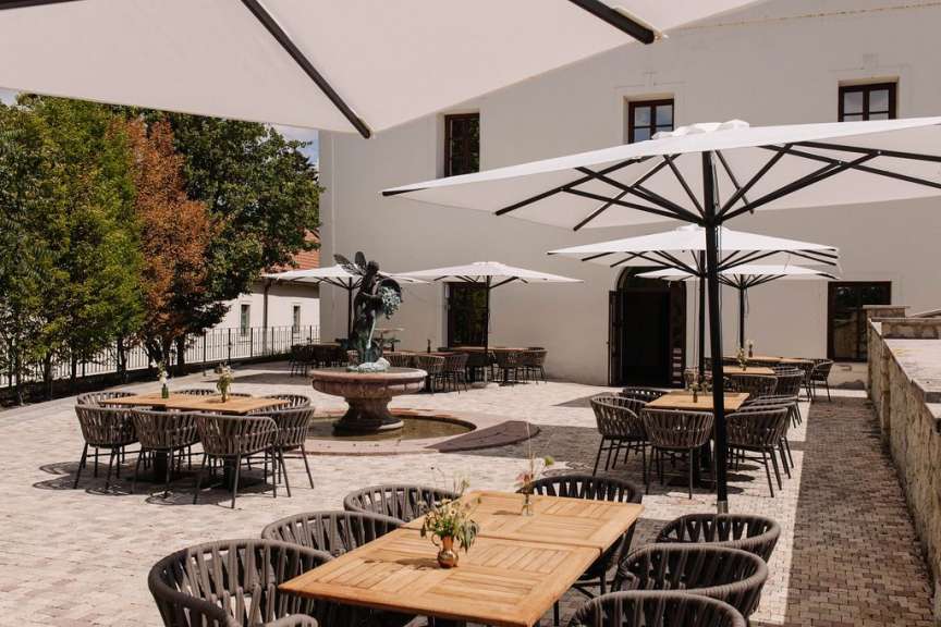 Die Aufnahme zeigt einen belebten Außenbereich eines Restaurants mit vielen Tischen, Stühlen und Sonnenschirmen vor einem weißen Gebäude mit einer Statue und einem Brunnen.