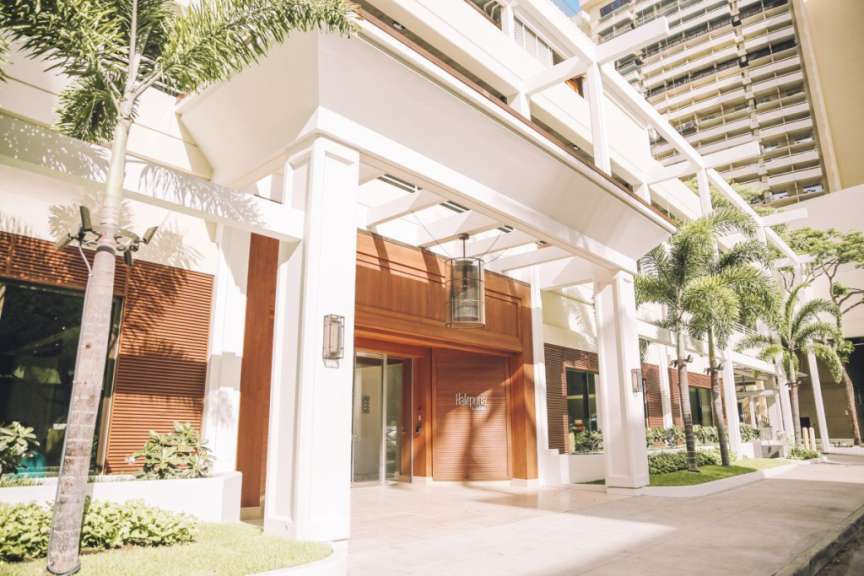 Die Eingangspartie eines modernen Hotels mit einem Vordach und Palmen im Vordergrund.