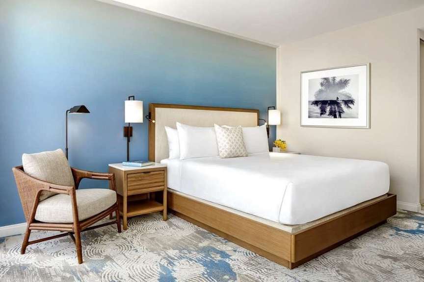 Ein modernes Schlafzimmer mit einem großen Bett, einem Sessel und einem Nachttisch wird von einem ombré-blauen Wandgemälde und einem gerahmten Bild mit Palmen und einem Vogel an der Wand eingerahmt.