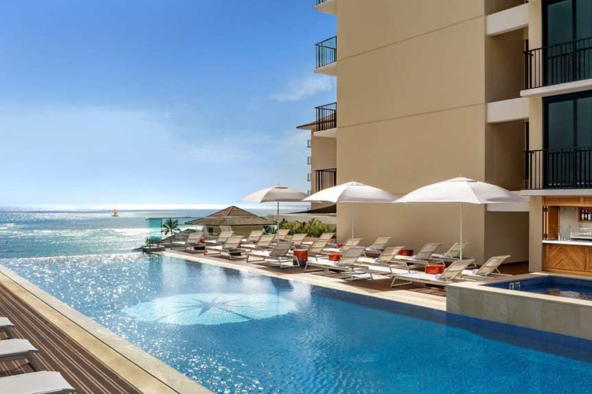 Ein Infinity-Pool mit Liegestühlen und Sonnenschirmen ist vor einem Hotelgebäude und dem Meer zu sehen.