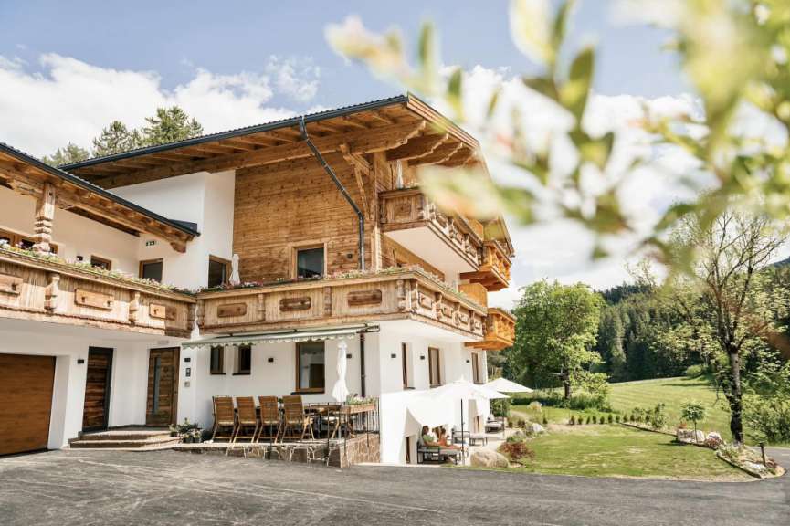 Ein Holzhaus mit Balkonen, Gäste, die auf einer Terrasse sitzen, und ein großer Garten mit Bäumen und einer grünen Wiese.