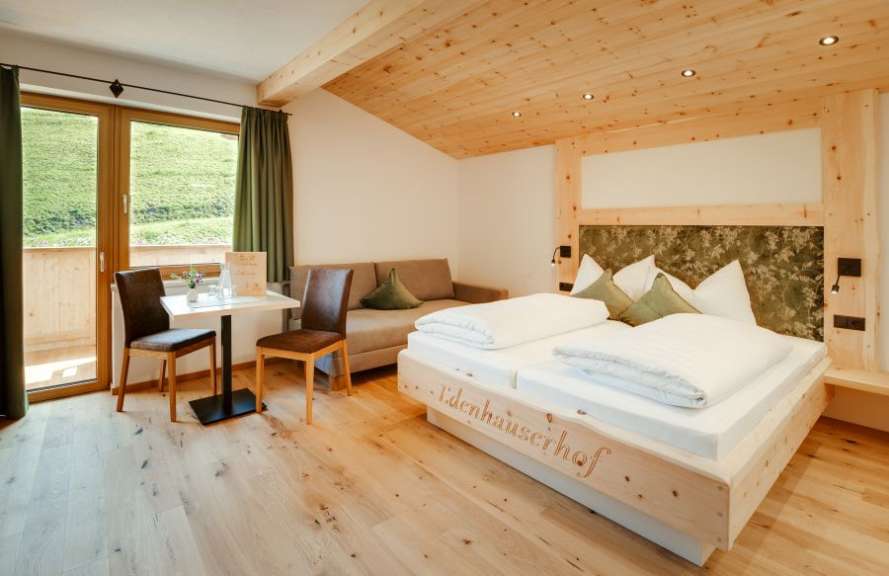 Ein geräumiges Hotelzimmer mit einem Doppelbett, einem Sofa, einem kleinen Tisch mit Stühlen und bodentiefen Fenstern, die auf eine grüne Landschaft blicken.