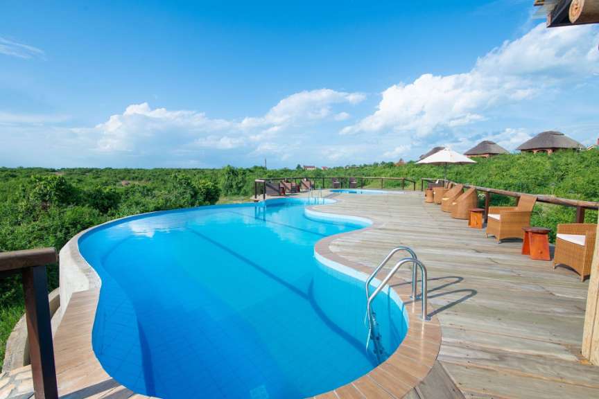 Ein gewundener Pool und eine Holzterrasse mit Sitzmöbeln sind von üppiger grüner Vegetation umgeben, während im Hintergrund strohgedeckte Gebäude und ein blauer Himmel mit Wolken zu sehen sind.