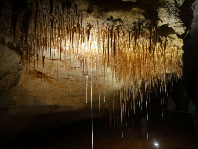 In einer Höhle sind viele Stalaktiten, beleuchtet von einem warmen Licht, zu sehen, während im Hintergrund Besucher auf einem Steg stehen.