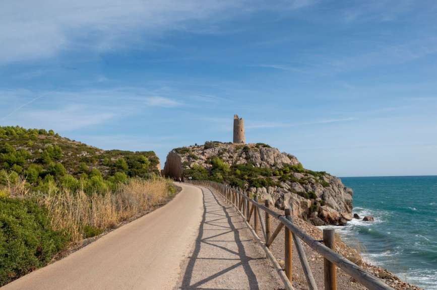 Ein Weg führt an einem felsigen Küstenabschnitt mit einem Wachturm auf einem Hügel entlang, der vom blauen Meer umgeben ist.