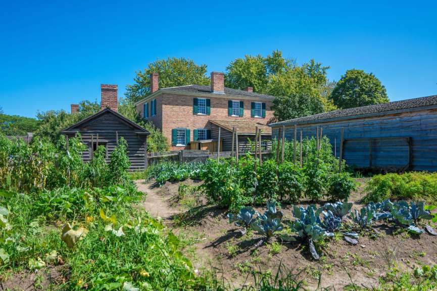 Ein Ziegelhaus und ein Holzschuppen stehen in einer ländlichen Umgebung mit einem Gemüsegarten im Vordergrund.