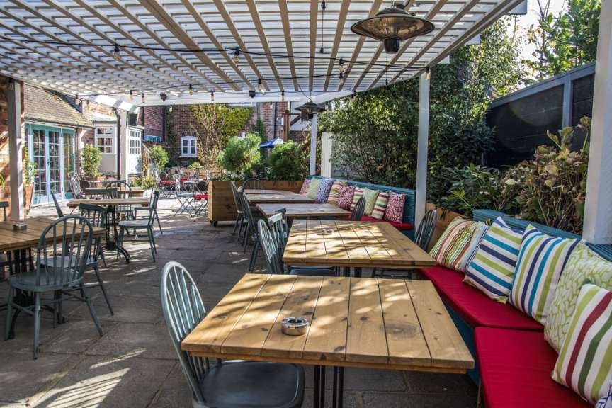 Auf der Terrasse eines Restaurants stehen Holztische mit Stühlen, bedeckt von einer Pergola mit Lichtern und einem Heizstrahler.