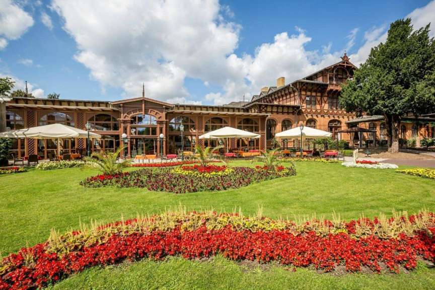 Ein großes, historisches Gebäude aus Holz und Backstein mit vielen Fenstern und Balkonen ist von einem gepflegten Garten mit bunten Blumenbeeten und Rasenflächen umgeben.