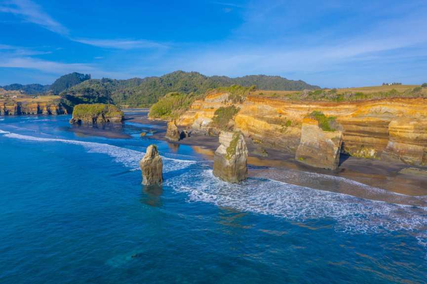 Eine Drohnenaufnahme zeigt eine dramatische Küstenlandschaft mit orangefarbenen Klippen, schwarzen Sandstränden, Felsnadeln im Meer und üppigem Grün im Hintergrund unter einem blauen Himmel.