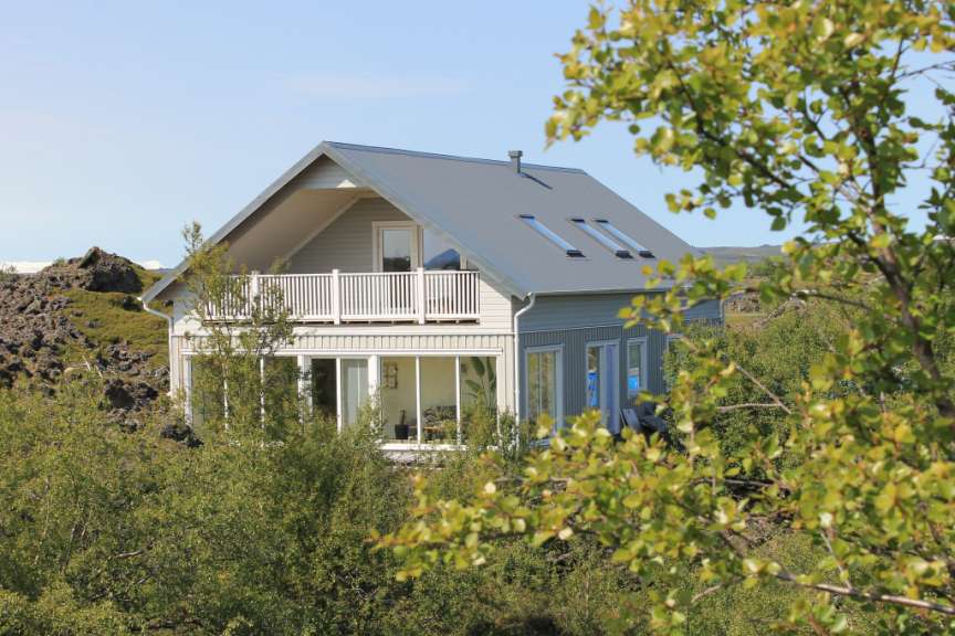 Ein Haus mit grauem Metalldach, weißem Balkon und großen Fenstern steht inmitten grüner Bäume und Felsen unter einem blauen Himmel.
