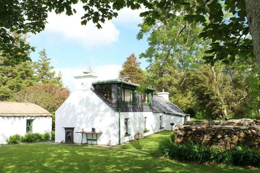 Ein weiß getünchtes Haus mit grünen Fensterrahmen steht auf einem sonnigen Rasen vor einem Wald.
