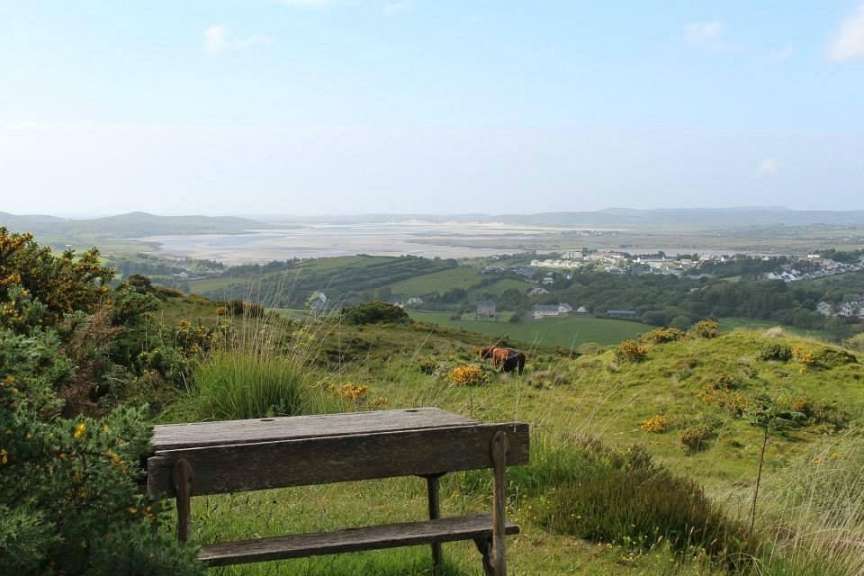 Ein hölzerner Sitzplatz bietet einen Ausblick auf eine Stadt, die in einer hügeligen Landschaft liegt, mit zwei Kühen auf den Weiden.