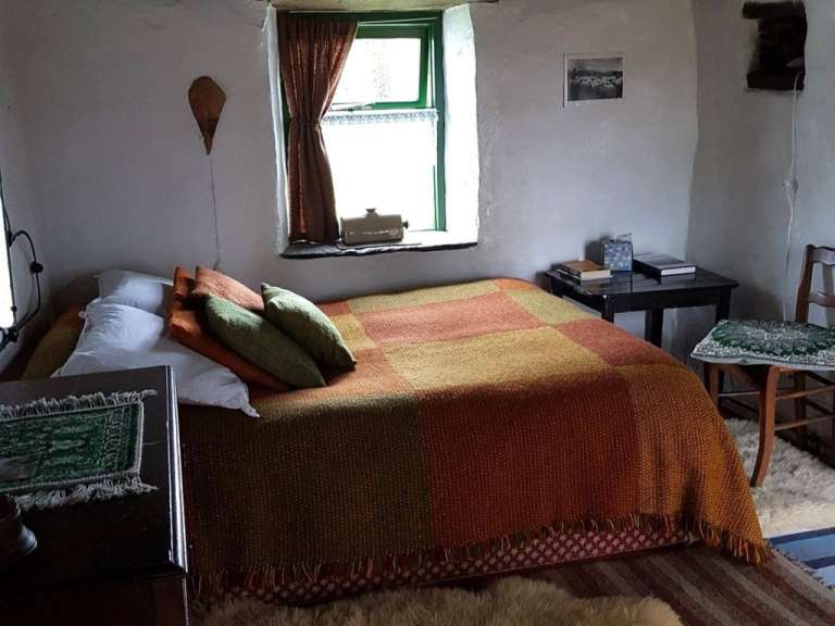 Das Bild zeigt ein Schlafzimmer mit einem Bett, das mit einer karierten Decke und Kissen bedeckt ist, einem Nachttisch mit Büchern und einer kleinen Schubladeneinheit.