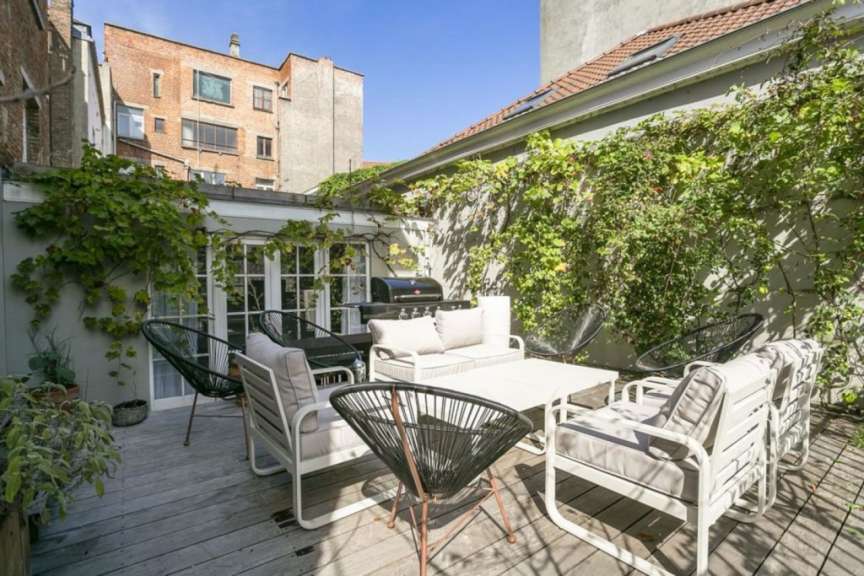 Ein sonniger Innenhof mit einer Sitzgruppe, einem Grill und einem Tisch ist von üppigem Grün umgeben.