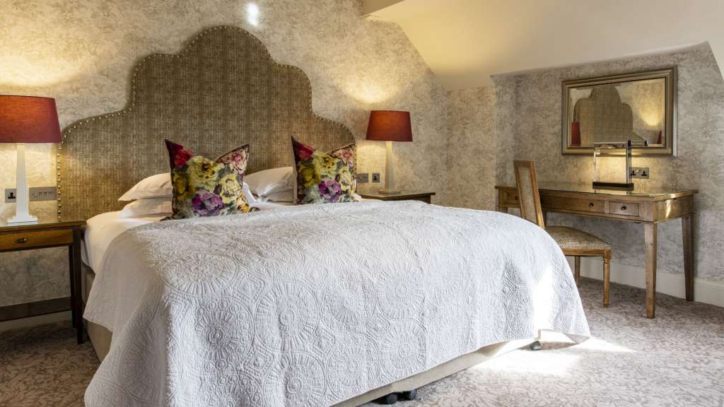 Ein bequemes Bett mit einer auffälligen Kopfteil, zwei dekorativen Kissen mit Blumenmuster und einer weißen Tagesdecke steht in einem gut beleuchteten Zimmer.